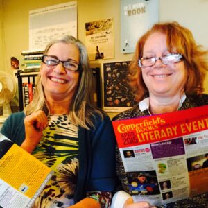Vicki DeArmon and Grace Bogart stand in front of a wall of literary posters and laugh together holding Copperfield's Books Event Calendars